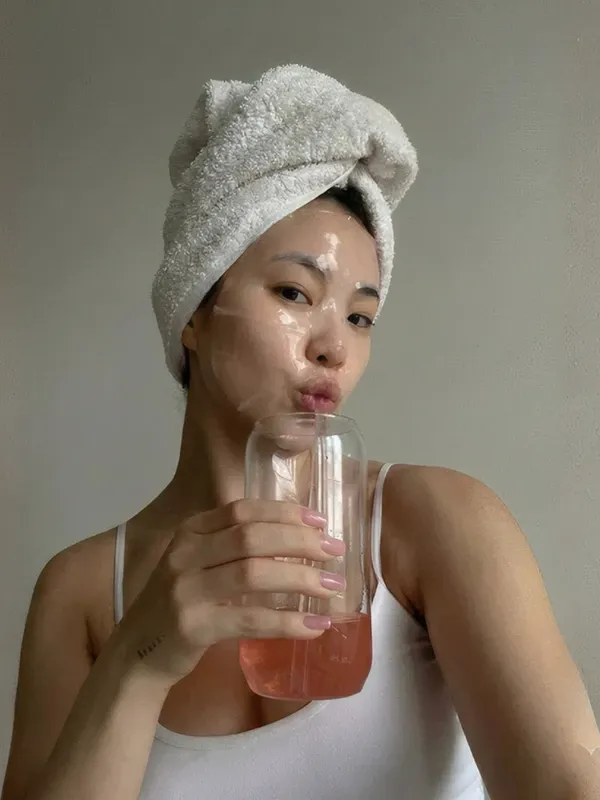 Una mujer con el cabello envuelto en una toalla y una mascarilla puesta, tomando un jugo en casa.